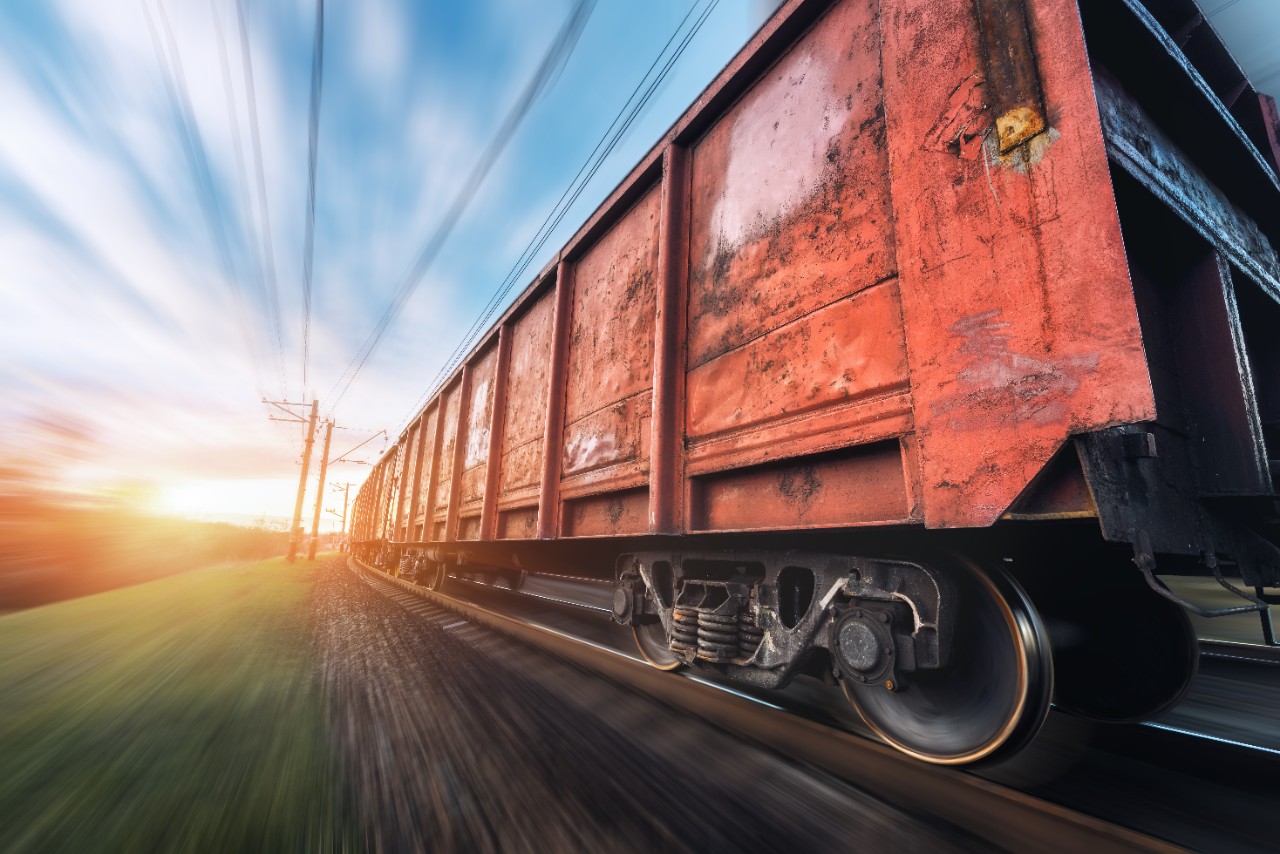 Railway station with cargo wagons and train in motion against sunny sky. Concept industrial landscape at sunset. Railroad. Motion blur effect. Blurred railway platform. Heavy industry. Cargo shipping